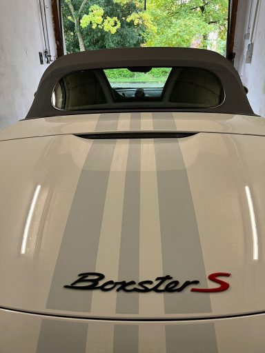 Weißes Auto mit grauen Streifen und "Boxster S"-Logo auf der Rückseite.