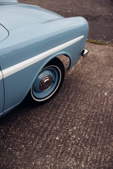 Blauer Oldtimer mit weißem Streifen und Stahlfelge auf einer rauen Fläche.