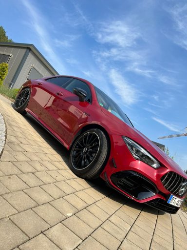 Roter Sportwagen mit schwarzen Felgen, auf einem gepflasterten Parkplatz unter blauem Himmel.
