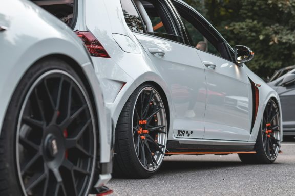Weißer Sportwagen mit tiefem Fahrwerk und schwarzen Felgen, im Fokus die Vorderachse.
